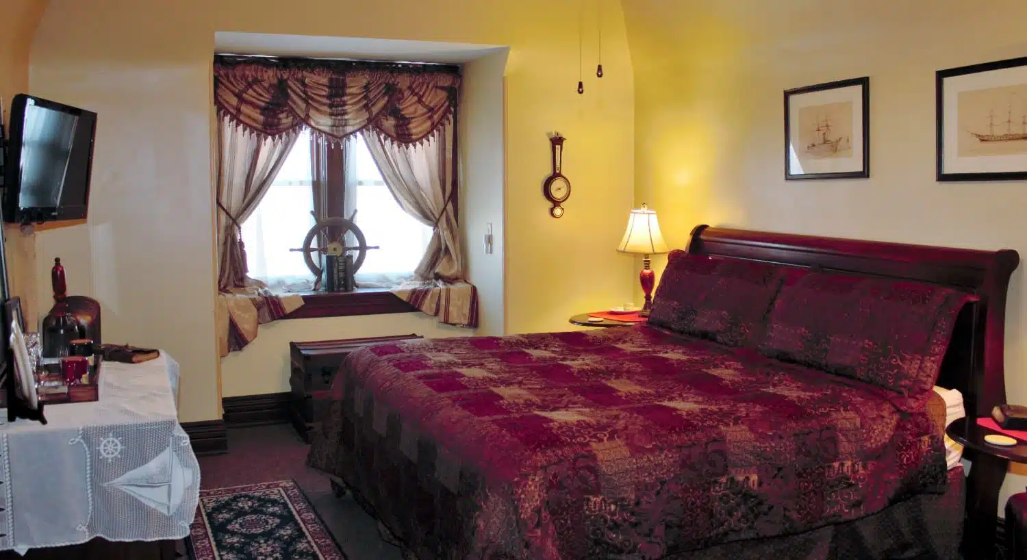 Bedroom with cream walls, dark brown trim, wooden sleigh bed, burgundy bedding, window nook, and wall-mounted TV