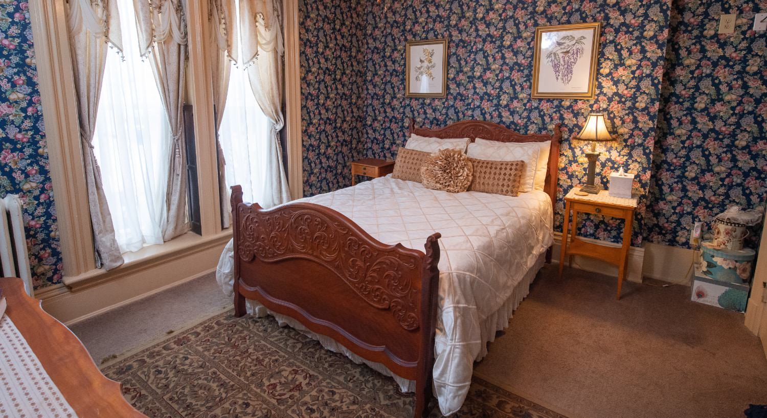 Bedroom with floral wallpaper, tan carpeting, wooden ornate bed, white bedding, wooden nightstands, and large draped windows