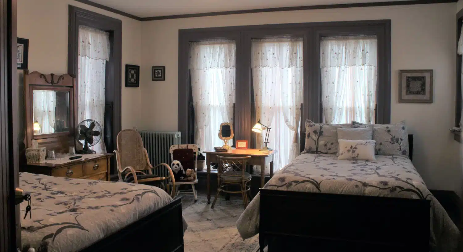 Large bedroom with white walls, gray trim, two dark wooden beds with multicolored bedding, antique wooden dresser, and small desk