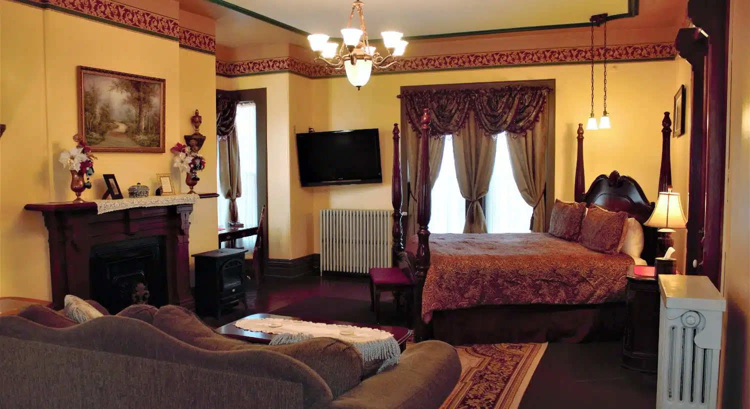 Large bedroom suite with yellow walls, hardwood flooring, four poster bed, multicolored burgundy paisley bedding, tan couch, fireplace, and wall-mounted TV