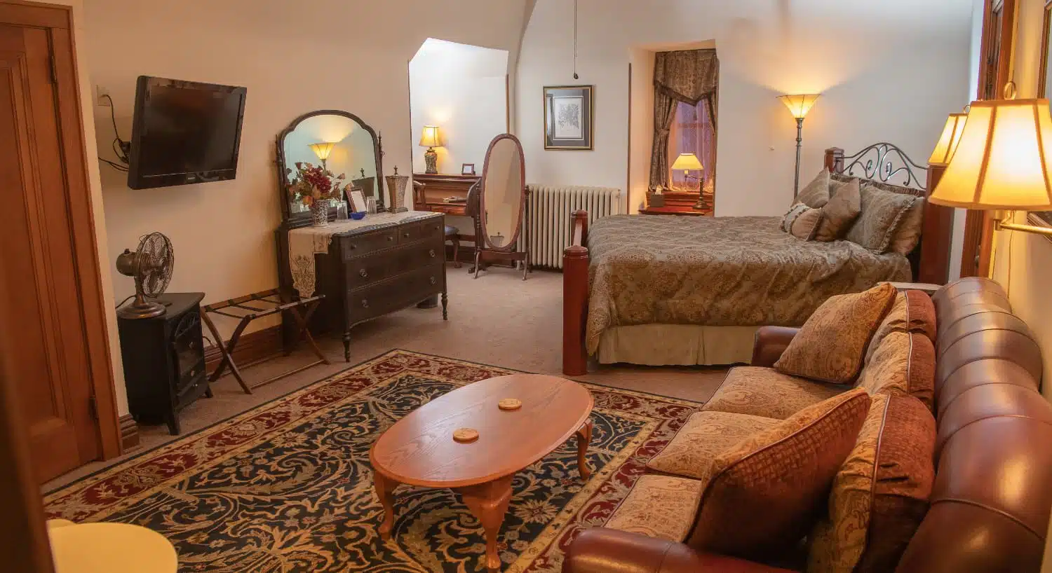 Large bedroom suite with white walls, tan carpeting, brown couch, wooden coffee table, antique dresser, and wall-mounted TV