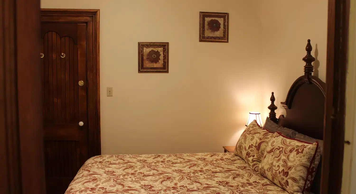 Bedroom with dark wooden vintage headboard, multicolored white and burgundy paisley bedding, and two small frames with a red flower hanging on the wall